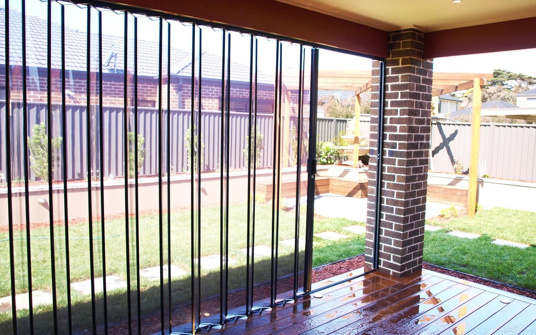 Clear folding outdoor blinds closed across a covered timber deck, rain on the boards and the garden visible through the panels